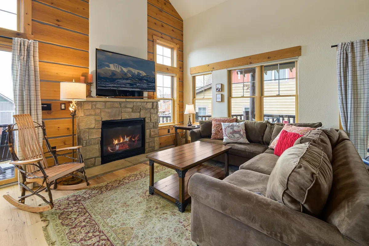 A cozy living room centered around a stone fireplace, with a plush sofa, rocking chair, coffee table, and a large television. Expansive windows let in mountain light, and the space exudes warmth and comfort—perfect for relaxing or gathering with loved ones.