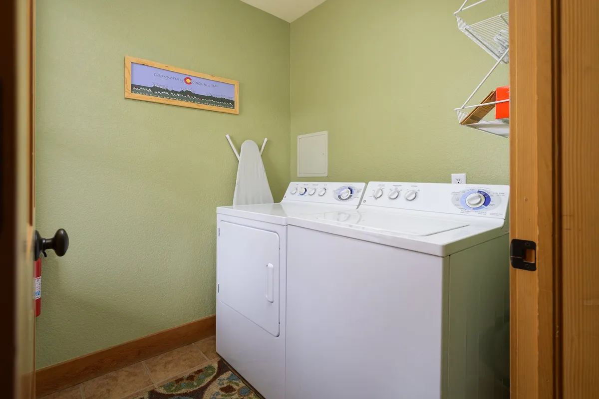 A tidy laundry area equipped with a washing machine, dryer, ironing board, and storage shelves with orange containers. The space is bright, organized, and ready for post-adventure clean-up.