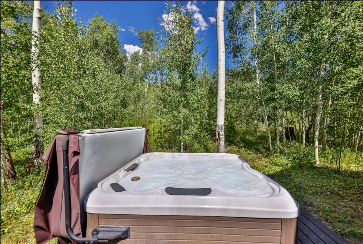 This image features a private outdoor hot tub surrounded by lush trees and a clear blue sky, creating a peaceful and secluded atmosphere. The hot tub is set on a wooden deck, suggesting a perfect spot to relax and enjoy nature.