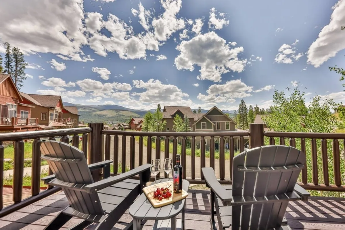 The image captures two Adirondack chairs with wine glasses and a fruit platter set on a deck, overlooking a breathtaking mountain view. The scene exemplifies serene outdoor leisure and socializing while enjoying nature.