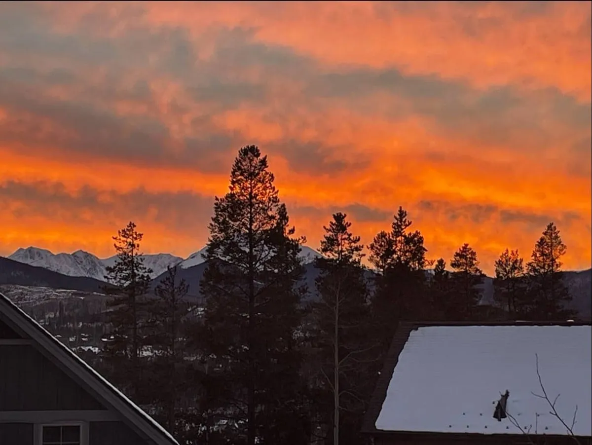 A tranquil exterior scene featuring a sweeping sunset over majestic mountain peaks, framed by treetops and rooftops. The soft golden light creates a serene atmosphere, perfect for unwinding outdoors. This visual captures the dramatic vistas that define luxury mountain living.