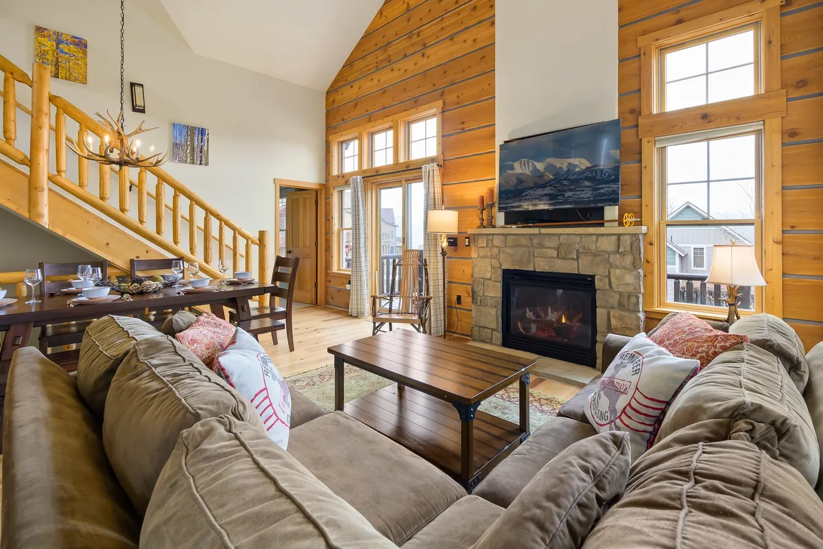 An open-concept interior showcasing a large sofa, a striking stone fireplace, a spacious dining table, a wooden staircase, and extra-large windows that frame scenic outdoor views. The layout is airy and bright, ideal for gatherings and shared moments.