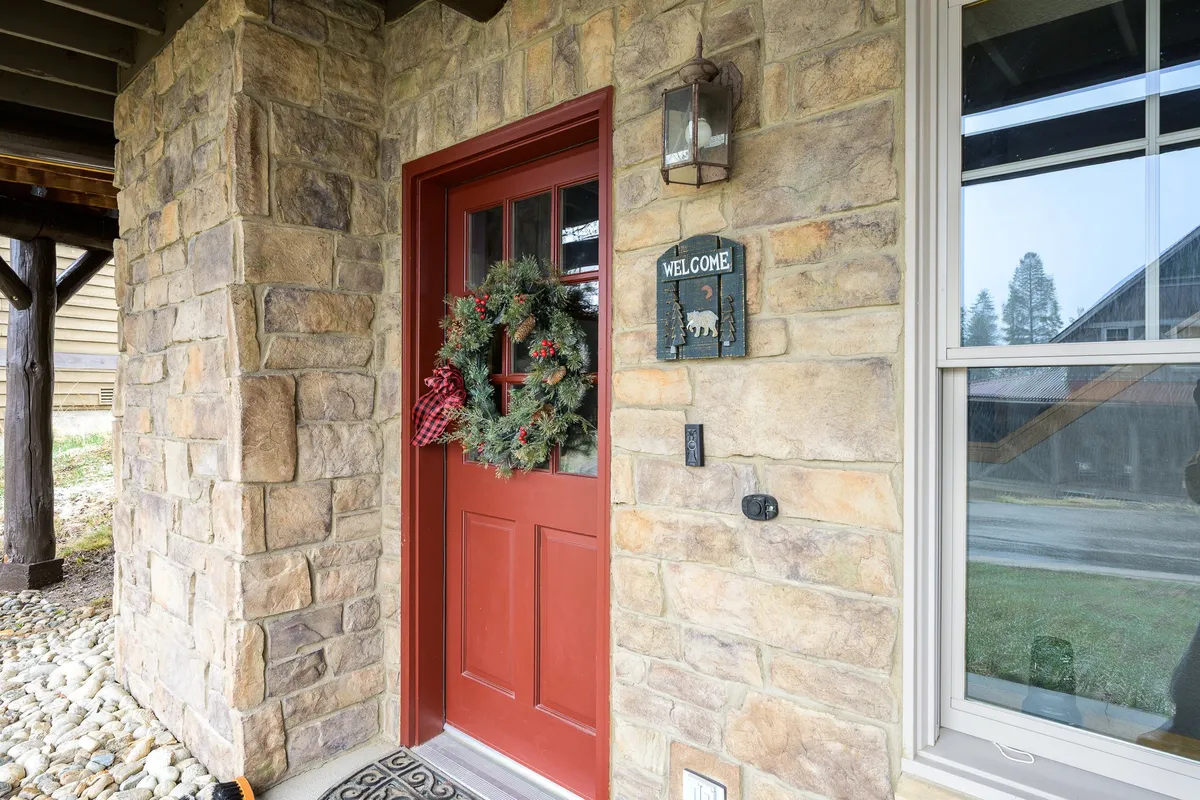 A close-up exterior of a welcoming stone facade with a bold red front door, festive wreath, and a friendly welcome sign. The cozy entrance is framed by natural stone and subtle greenery, embodying both hospitality and mountain charm.