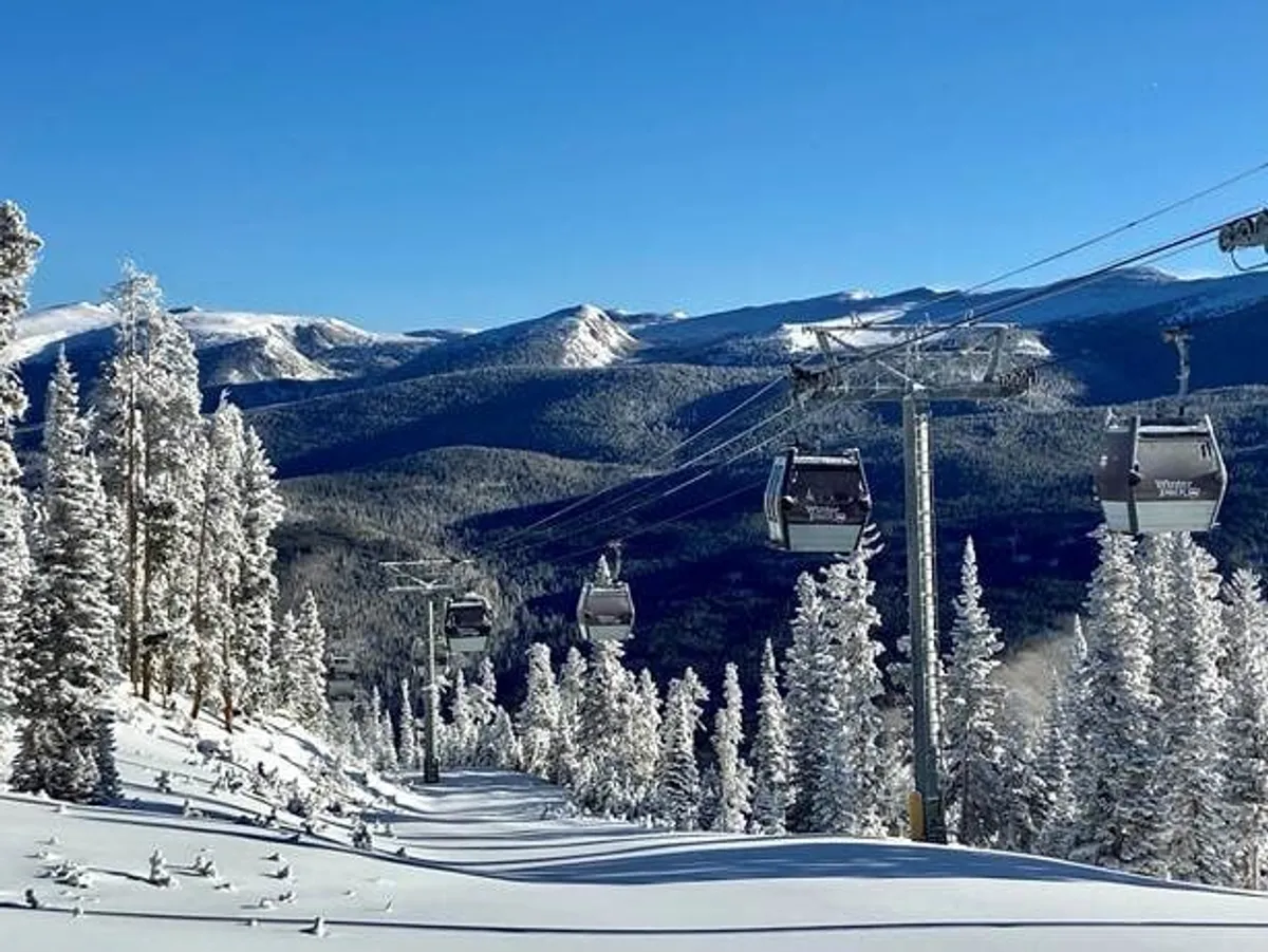 A dramatic ski lift rises above snow-covered trees with a mountain range in the background. Skiers and snowboarders can be seen descending on pristine powder, and expansive winter vistas stretch to the horizon.