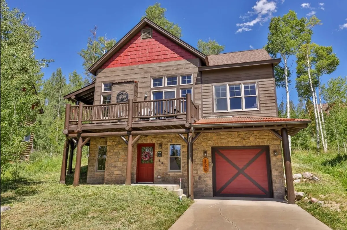The image features the exterior of a luxurious mountain cabin in Fraser, Colorado, showcasing a welcoming front porch with a bold red door, a striking stone facade, and an inviting wooden balcony. The setting hints at opportunities for relaxing on the porch, watching sunsets from the balcony, and enjoying the natural surroundings with family and friends.