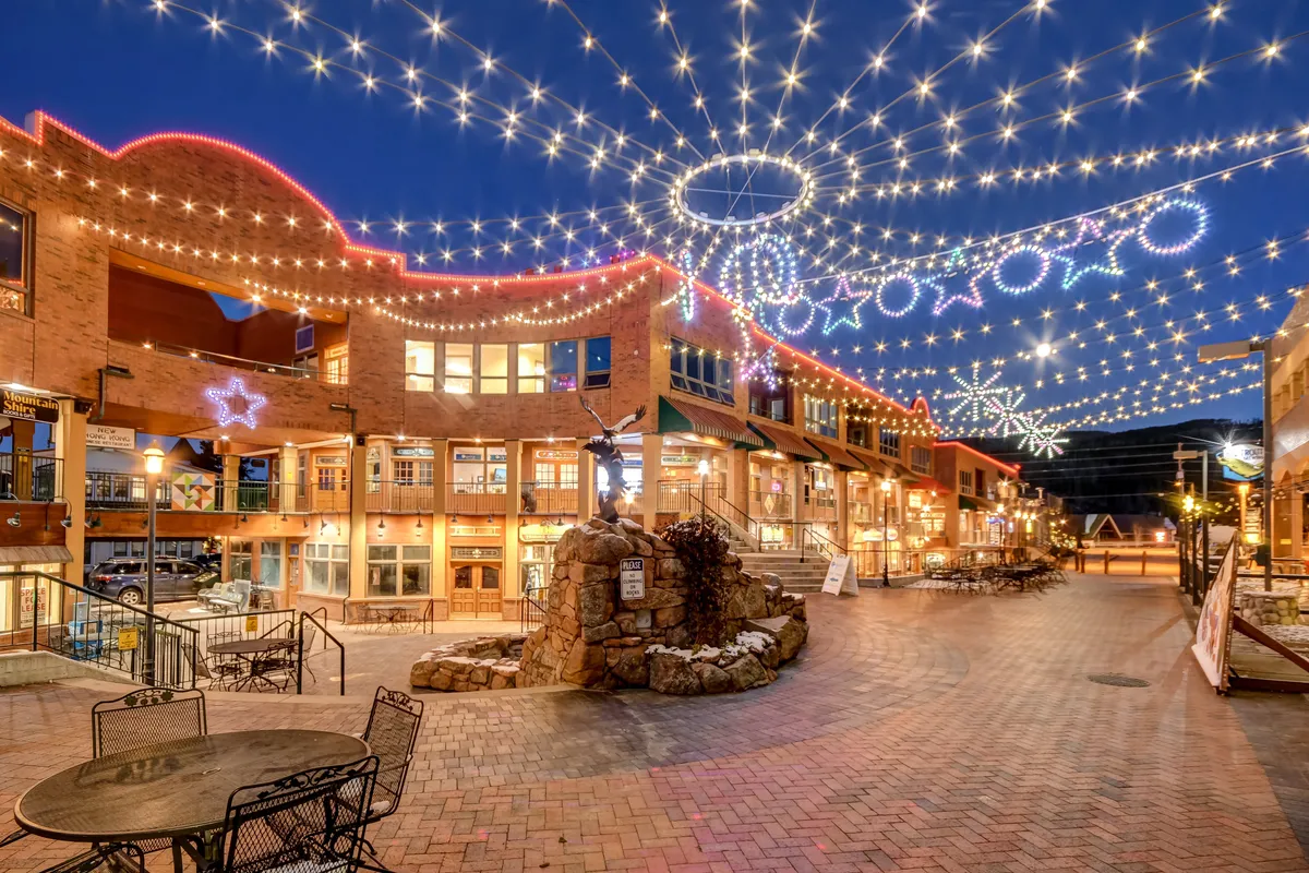 A lively outdoor plaza features string lights overhead, a charming stone fountain, and cozy outdoor seating areas surrounded by decorative storefronts. Guests are seen socializing, shopping, dining al fresco, and participating in festive community activities.