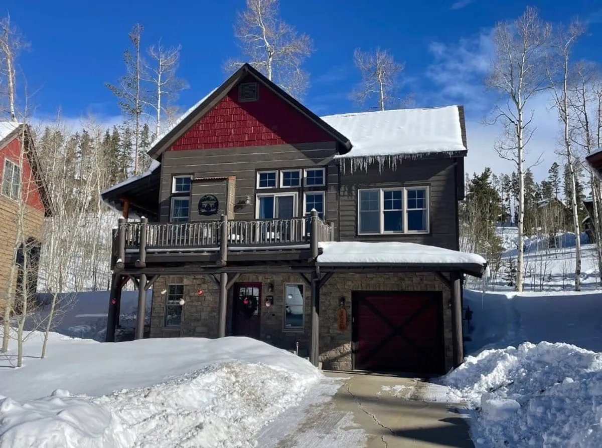 A luxury home exterior with a wooden balcony, stone facade, and red accents, set against a snowy driveway. The scene hints at family gatherings, relaxing on the balcony, and winter sports right outside the door.