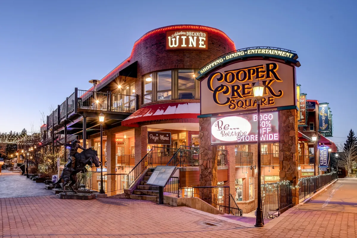 This inviting shot showcases the Cooper Creek Square sign, wine tasting signage, outdoor dining spaces, and a horse sculpture, bustling with patrons enjoying shopping, dining, and entertainment options in downtown Winter Park.