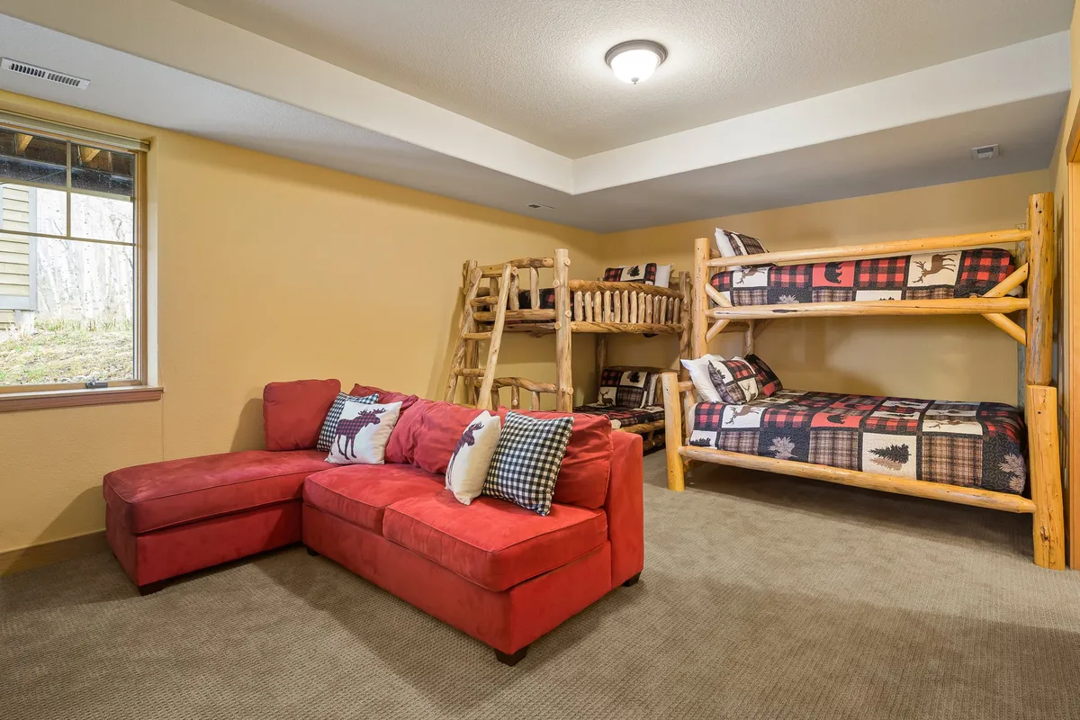 A playful, kid-friendly bedroom featuring bunk beds with plaid bedding, a red sectional sofa, and a window letting in natural light. The arrangement includes seating for games and reading, perfect for group or family stays.